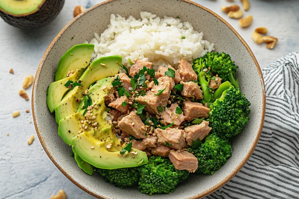 Fresh ingredients for Broccoli Avocado Tuna Bowl, including broccoli, avocado, tuna, lemon, and olive oil on a marble countertop.